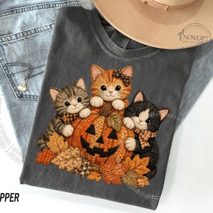 Könnte beinhalten: Ein anthrazitfarbenes T-Shirt mit einer Grafik von drei Kätzchen, die um einen geschnitzten orangefarbenen Kürbis sitzen, der von Herbstblättern umgeben ist. Die Kätzchen tragen kleine gemusterte Schleifen. Das T-Shirt liegt auf einer blauen Jeans und einem beigen Hut.