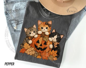 Op de afbeelding: Een antracietgrijs t-shirt met een afbeelding van drie kittens die rond een uitgesneden oranje pompoen zitten, omringd door herfstbladeren. De kittens dragen kleine gestreepte strikjes. Het t-shirt wordt getoond met een blauwe spijkerbroek en een beige hoed.