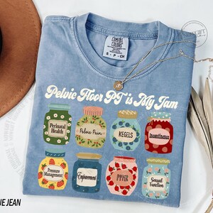 May include: A light blue t-shirt with the text "Pelvic Floor PT is My Jam" and illustrations of jars labeled with terms like "Perinatal Health" and "Kegels." A gold necklace and a brown hat are also visible.