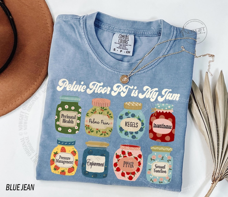 May include: A light blue t-shirt with the text "Pelvic Floor PT is My Jam" and illustrations of jars labeled with terms like "Perinatal Health" and "Kegels." A gold necklace and a brown hat are also visible.