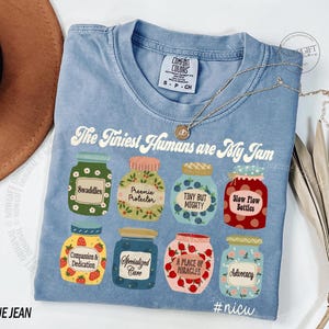 Puede incluir: Una camiseta azul claro con el texto "The Tiniest Humans are My Jam" e ilustraciones de tarros. Los tarros tienen etiquetas como "Swaddles" y "Advocacy". Un collar dorado est&aacute; drapeado sobre la camiseta. La camiseta est&aacute; etiquetada "BLUE JEAN".