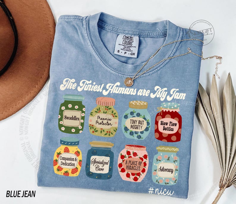 Puede incluir: Una camiseta azul claro con el texto "The Tiniest Humans are My Jam" e ilustraciones de tarros. Los tarros tienen etiquetas como "Swaddles" y "Advocacy". Un collar dorado est&aacute; drapeado sobre la camiseta. La camiseta est&aacute; etiquetada "BLUE JEAN".