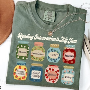 May include: Green t-shirt with the text "Reading Intervention is My Jam" and illustrations of jars with different reading skills, such as phonics, comprehension, fluency, vocabulary, phonemic awareness, content knowledge, language, and writing.