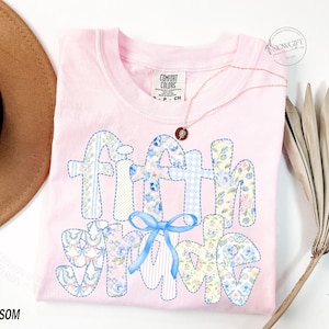 May include: A light pink Comfort Colors t-shirt with the words "Faith Shine" appliqued in various floral and patterned fabrics. A light blue bow is centered below the words. A gold necklace with a pendant is on the shirt. The word "Blossom" is at the bottom left.