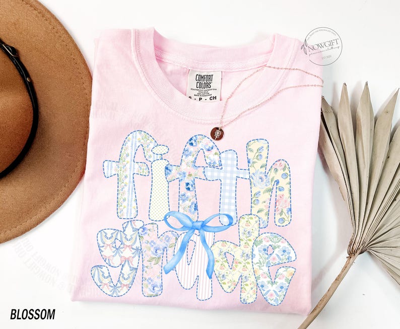 May include: A light pink Comfort Colors t-shirt with the words "Faith Shine" appliqued in various floral and patterned fabrics. A light blue bow is centered below the words. A gold necklace with a pendant is on the shirt. The word "Blossom" is at the bottom left.
