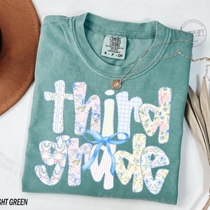 Puede incluir: Camiseta verde claro Comfort Colors con las palabras "Third Grade" aplicadas en telas florales y estampadas. Un lazo azul claro est&aacute; centrado debajo de la palabra "Third". Un collar dorado con un colgante descansa sobre la camisa.