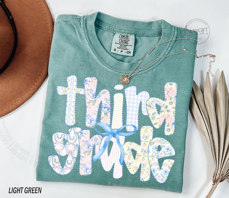 Puede incluir: Camiseta verde claro Comfort Colors con las palabras "Third Grade" aplicadas en telas florales y estampadas. Un lazo azul claro est&aacute; centrado debajo de la palabra "Third". Un collar dorado con un colgante descansa sobre la camisa.
