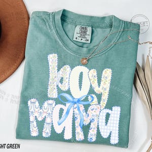 Puede incluir: Camiseta verde claro con las palabras "Joy Mama" en un dise&ntilde;o de patchwork. Las letras son blancas, florales y de cuadros azules. Un lazo azul claro est&aacute; entre las palabras. Un collar dorado con un colgante est&aacute; en la camiseta.