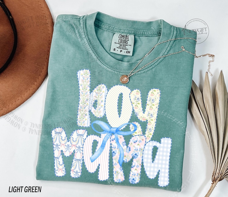 Puede incluir: Camiseta verde claro con las palabras "Joy Mama" en un dise&ntilde;o de patchwork. Las letras son blancas, florales y de cuadros azules. Un lazo azul claro est&aacute; entre las palabras. Un collar dorado con un colgante est&aacute; en la camiseta.