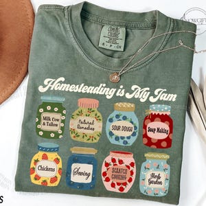 May include: Moss green t-shirt with the phrase "Homesteading is My Jam" and illustrations of jars labeled with homesteading activities. A gold necklace is draped across the shirt. The shirt is made of Comfort Colors material.