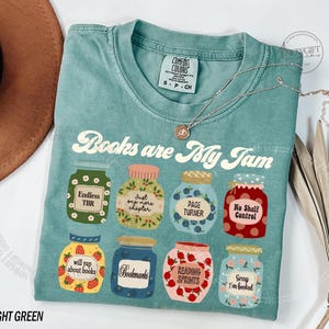 May include: A light green t-shirt featuring the text "Books are My Jam" and various jam jar illustrations. Each jar has a different label, including "Endless TBR" and "Bookmarks". A silver necklace with a small pendant is also visible.