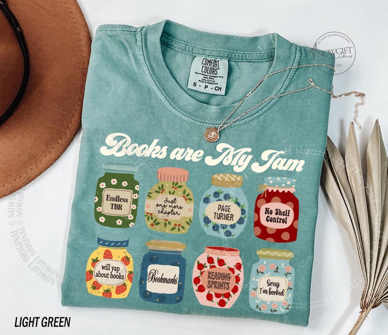 May include: A light green t-shirt featuring the text "Books are My Jam" and various jam jar illustrations. Each jar has a different label, including "Endless TBR" and "Bookmarks". A silver necklace with a small pendant is also visible.