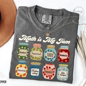 May include: Gray t-shirt with the text "Math is My Jam" in a retro font. The shirt features a design of eight jars with different math-related terms, such as "Numbers", "Measurement", "Algebraic Thinking", and "Geometry".