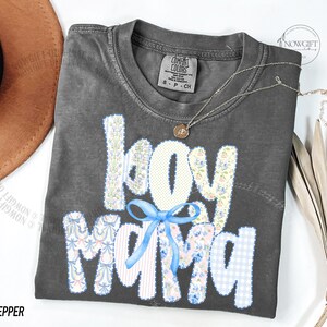 Puede incluir: Una camiseta gris oscuro con las palabras "boy mama" en un dise&ntilde;o de patchwork. Las letras son blancas, florales y de cuadros azules. Un lazo azul claro est&aacute; centrado debajo de la palabra "boy". Un collar plateado con un peque&ntilde;o colgante est&aacute; en la camisa.