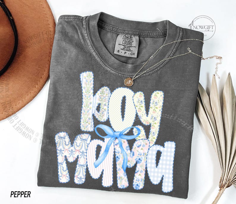 Puede incluir: Una camiseta gris oscuro con las palabras "boy mama" en un dise&ntilde;o de patchwork. Las letras son blancas, florales y de cuadros azules. Un lazo azul claro est&aacute; centrado debajo de la palabra "boy". Un collar plateado con un peque&ntilde;o colgante est&aacute; en la camisa.