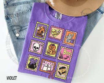 May include: A violet-colored t-shirt featuring a Halloween-themed stamp design. The stamps include images of a haunted house, a heart with text, a mummy, a skull, pumpkins, a witch's cauldron, ghosts, and a bat. The shirt is paired with blue jeans and a hat.