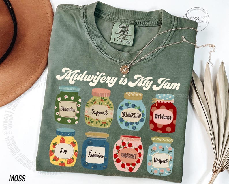 May include: Moss green t-shirt with the phrase "Midwifery is My Jam" and jars labeled "Education," "Support," "Collaboration," "Evidence," "Joy," "Inclusion," "Consent," and "Respect." A silver necklace is draped across the shirt.