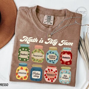 May include: A brown t-shirt with the text "Math is My Jam" printed on it. The text is in a retro font. There are nine jars with different math-related terms printed on them, such as "Numbers", "Measurement", "Algebraic Thinking", "Geometry", "Operations", "Problem Solving", "Probability", and "Data Analysis".