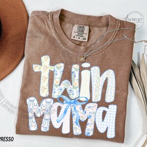 Puede incluir: Camiseta marr&oacute;n con la frase "twin mama" en un dise&ntilde;o de patchwork. Las letras presentan estampados florales, rayas y cuadros vichy. Un lazo azul adorna el dise&ntilde;o. Un collar plateado con un colgante est&aacute; sobre la camiseta.