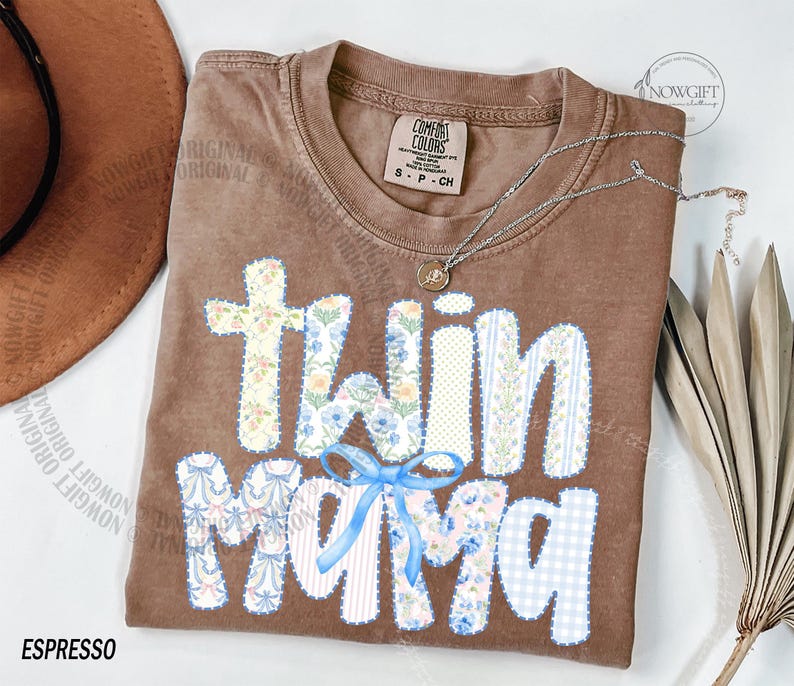 Puede incluir: Camiseta marr&oacute;n con la frase "twin mama" en un dise&ntilde;o de patchwork. Las letras presentan estampados florales, rayas y cuadros vichy. Un lazo azul adorna el dise&ntilde;o. Un collar plateado con un colgante est&aacute; sobre la camiseta.