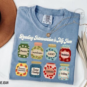 May include: A blue t-shirt with the text "Reading Intervention is My Jam" printed on it. The text is surrounded by illustrations of jars with different reading skills listed on them, such as "phonics", "comprehension", "fluency", and "vocabulary".