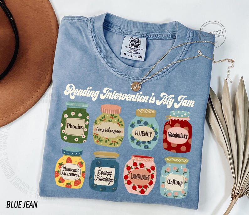 May include: A blue t-shirt with the text "Reading Intervention is My Jam" printed on it. The text is surrounded by illustrations of jars with different reading skills listed on them, such as "phonics", "comprehension", "fluency", and "vocabulary".