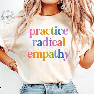 May include: Ivory-colored t-shirt with the words "practice radical empathy" in colorful, bold letters. The shirt has short sleeves and a relaxed fit. The text is in a rainbow of colors: pink, blue, red, orange, and purple.