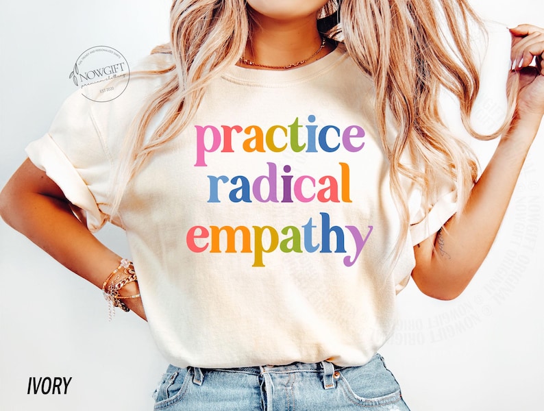 May include: Ivory-colored t-shirt with the words "practice radical empathy" in colorful, bold letters. The shirt has short sleeves and a relaxed fit. The text is in a rainbow of colors: pink, blue, red, orange, and purple.