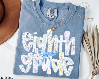 Op de afbeelding: Blauwe spijkerbroek Comfort Colors T-shirt met de woorden "Eighth Grade" in een patchwork ontwerp. De letters zijn gemaakt van bloemen- en patronenstoffen. Een blauwe strik staat onder de tekst en een gouden ketting ligt op het shirt.