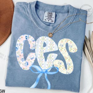 May include: A blue jean Comfort Colors t-shirt with a floral design spelling out the word "CPS". The letters are decorated with a mix of floral patterns and a blue bow at the bottom. A gold necklace with a pendant is draped across the shirt.