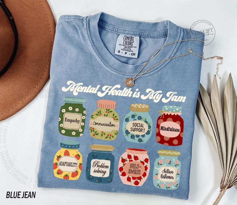 K&ouml;nnte beinhalten: Ein blaues Jeans-T-Shirt mit dem Aufdruck "Mental Health is My Jam" auf der Vorderseite. Der Text ist in einer Retro-Schriftart gehalten und von Illustrationen von Gl&auml;sern mit verschiedenen mentalen Gesundheitskonzepten umgeben, wie z. B. "Empathie", "Kommunikation", "Soziale Unterst&uuml;tzung", "Achtsamkeit", "Anpassungsf&auml;higkeit", "Probleml&ouml;sung", "Selbstwahrnehmung" und "Aktives Zuh&ouml;ren".