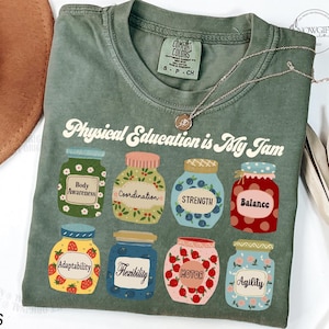 May include: A green t-shirt with the text "Physical Education is My Jam" in a retro font. The shirt features a design of nine jars with different aspects of physical education, such as "Body Awareness", "Coordination", "Strength", "Balance", "Adaptability", "Flexibility", "Motor", and "Agility".