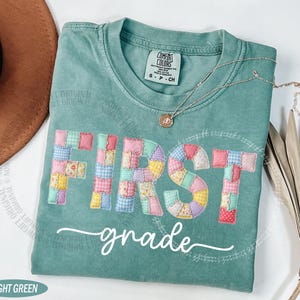 Peut inclure: T-shirt vert clair avec l'inscription "FIRST grade" dans un motif patchwork. Le t-shirt a un col rond et une petite étiquette en haut à gauche. Un collier argenté avec un pendentif est drapé sur le t-shirt.