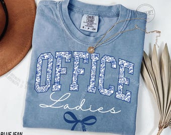 Op de afbeelding: Een lichtblauw t-shirt met de woorden "OFFICE Ladies" in witte letters en een blauw bloemenpatroon. Een gouden ketting met een hanger is over het shirt gedrapeerd. Het shirt heeft een klein label met "Comfort Colors".