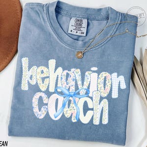 Könnte beinhalten: Blaues Comfort Colors T-Shirt mit dem Schriftzug "behavior coach" in weißen Buchstaben mit floralem Muster. Eine hellblaue Schleife befindet sich zwischen den Wörtern. Eine goldene Halskette mit Anhänger befindet sich auf dem Shirt.
