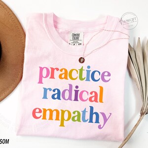 May include: A light pink t-shirt with the words "practice radical empathy" in rainbow colors. The shirt is a Comfort Colors brand. A gold necklace with a pendant is draped on the shirt. A brown hat and dried palm leaf are in the background.
