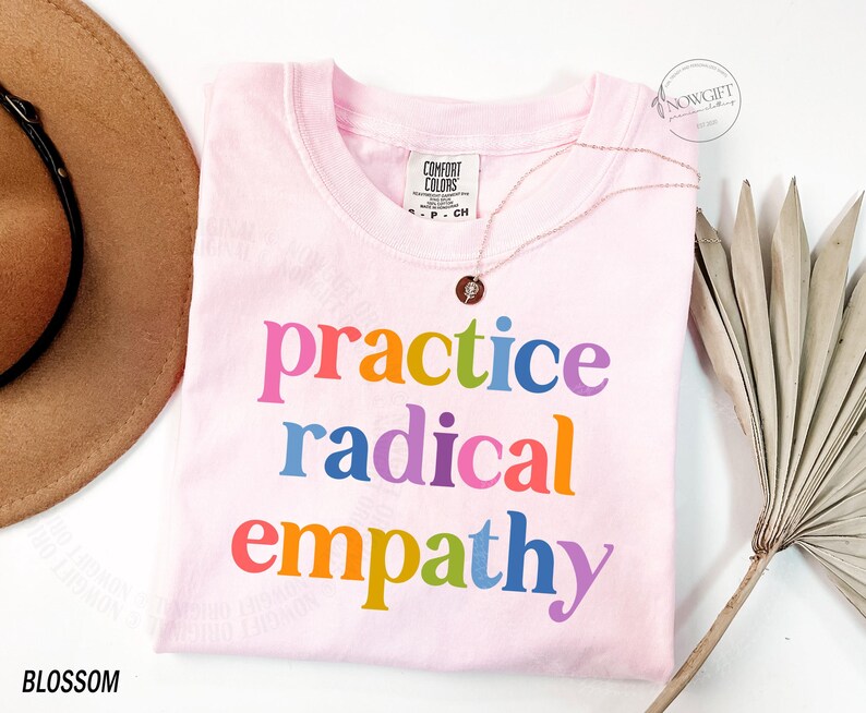 May include: A light pink t-shirt with the words "practice radical empathy" in rainbow colors. The shirt is a Comfort Colors brand. A gold necklace with a pendant is draped on the shirt. A brown hat and dried palm leaf are in the background.