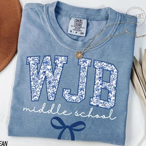 May include: A blue jean coloured t-shirt featuring the letters "WJB" in a floral design, with the words "middle school" in white script. A blue bow graphic is at the bottom. A gold necklace is draped across the shirt.