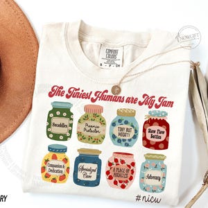 Puede incluir: Camiseta de color marfil con la frase "The Tiniest Humans are My Jam". La camiseta presenta ilustraciones de ocho tarros de mermelada coloridos, cada uno etiquetado con un t&eacute;rmino diferente. Un collar de oro est&aacute; drapeado sobre la camiseta.