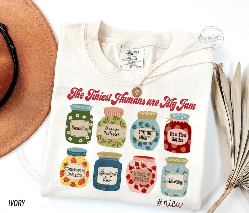 Puede incluir: Camiseta de color marfil con la frase "The Tiniest Humans are My Jam". La camiseta presenta ilustraciones de ocho tarros de mermelada coloridos, cada uno etiquetado con un t&eacute;rmino diferente. Un collar de oro est&aacute; drapeado sobre la camiseta.