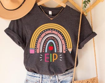 Op de afbeelding: Donkergrijs t-shirt met een kleurrijk regenboogontwerp, een rode appel en de letters "EIP". De regenboog heeft gele, roze, witte en blauwe bogen. Het shirt hangt aan een houten hanger.