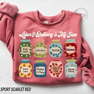 May include: A heather sport scarlet red sweatshirt with a graphic of jars of jam, each labeled with a stage of labor and delivery. The text "Labor & Delivery is My Jam" is printed in a retro font above the jars.