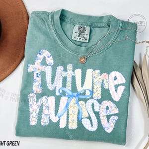May include: Light green t-shirt with the words "A Future Nurse" in a patchwork design. The letters are white with floral and gingham patterns. A light blue bow is between the words "Future" and "Nurse". A silver necklace with a small pendant is on the shirt.