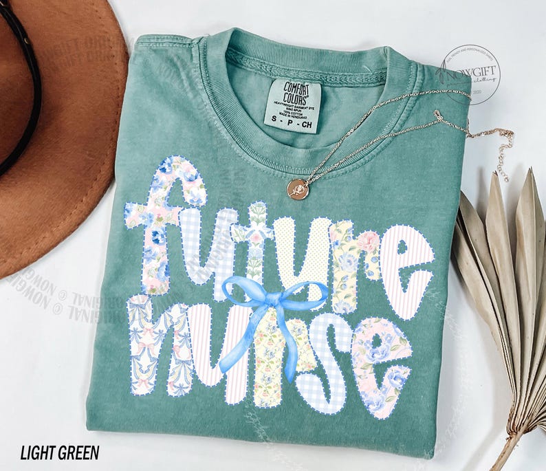 May include: Light green t-shirt with the words "A Future Nurse" in a patchwork design. The letters are white with floral and gingham patterns. A light blue bow is between the words "Future" and "Nurse". A silver necklace with a small pendant is on the shirt.
