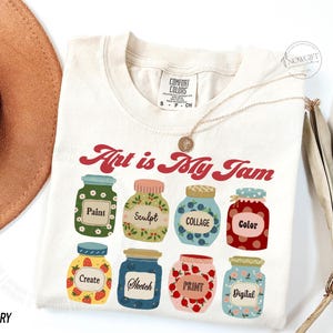 Op de afbeelding: Een ivoorwit T-shirt met de tekst "Art is My Jam" in rood en wit. Het shirt heeft een ontwerp van negen kleurrijke potten met verschillende kunstgerelateerde woorden erop, waaronder "Verf", "Beeldhouwen", "Collage", "Kleur", "Cre&euml;ren", "Schetsen", "Drukken" en "Digitaal".