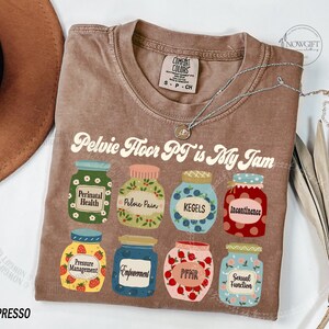 May include: Espresso-colored t-shirt with the text "Pelvic Floor PT is My Jam" and illustrations of jam jars. Each jar has a label with a different term related to pelvic floor health, such as "Perinatal Health" and "Kegels."