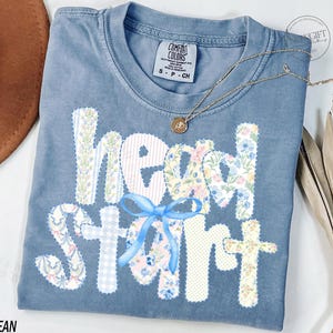 May include: A blue jean Comfort Colors t-shirt with the words "Head Start" appliqued in floral and patterned fabrics. A blue bow is centered on the shirt. A gold necklace with a pendant is on the shirt.