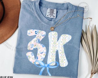 Op de afbeelding: Blauw spijkerbroek Comfort Colors T-shirt met een bloemen 3K-ontwerp en een blauwe strik. Het shirt heeft een klein label met de tekst "Comfort Colors" en "S-P-CH". Een gouden ketting met een hanger ligt op het shirt.