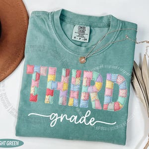 May include: Light green t-shirt with the words "THIRD grade" in a patchwork design. The shirt has a crew neck and short sleeves. A silver necklace with a pendant is draped on the shirt.