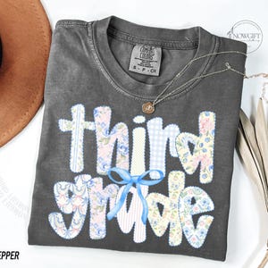 Puede incluir: Una camiseta gris Comfort Colors con las palabras "Third Grade" en un dise&ntilde;o de patchwork. Las letras est&aacute;n en patrones florales, de cuadros y a rayas, con un lazo azul. Tambi&eacute;n se ven un collar dorado y un sombrero marr&oacute;n.